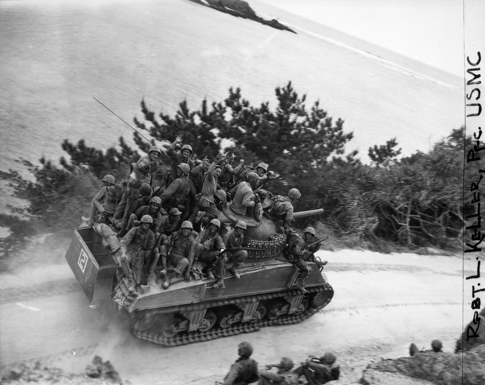 Riflemen of the 29th Marine Regiment ride a M4A3 Sherman 105mm of Company A, 6th Tank Battalion during the 6th Marine Division's drive on Chuda along the west coast of Okinawa. After expecting a contested landing on April 1, 1945, and seeing little of the Japanese, the Americans were in high spirits as objectives are taken ahead of schedule in Northern Okinawa; the Shuri Line would rob them of their high morale. The 29th Marines cut off the Motobu Peninsula and seized Chuda at 1200 Hours on April 6, 1945. Tank-infantry teams encountered sporadic resistance during the initial invasion; most problems were from the Japanese blowing bridges as they retreated inland. Destruction of bridges had been inept; frequently only a span of the bridge had been blown or cracked. The engineers cut quick bypasses for the vehicles, repairing the broken spans later. 500 M4A3 Shermans with the M4 105mm gun were built in late 1944. Later versions of the 105mm Sherman had a more advanced horizontal volute spring suspension (HVSS) with wider tracks that allowed for a smoother ride. Note partially dismantled deep wading gear to allow the M4A3 to move through deep water during the landings a few days before. The M4A3 Sherman with the M4 105mm howitzer was not popular with the tankers, who preferred the M1 76mm high-velocity gun in case of tank-against-tank engagements. However, the 105mm-equipped Shermans were very popular with ground troops, who used tanks as mobile pillboxes, taking out Japanese positions with point-blank high explosive fire.